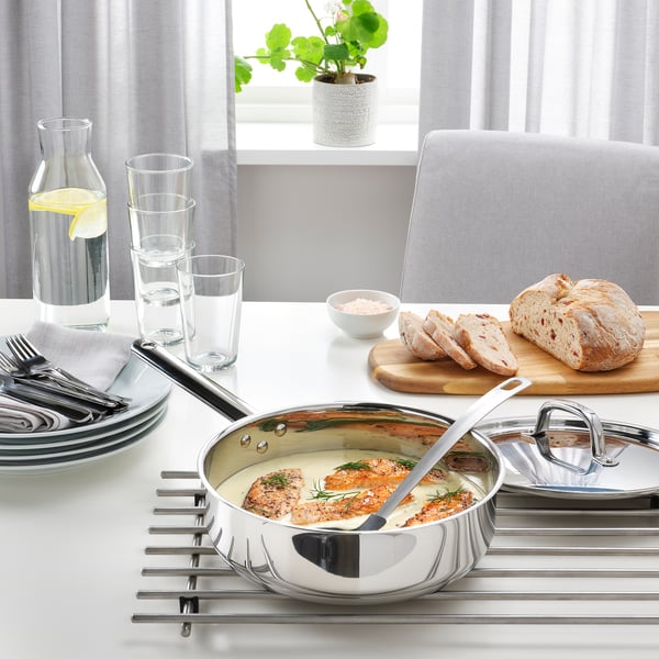 Sauterpande i rustfrit stål med låg på bordet, der indeholder tilberedt mad. Omgivet af tallerkener, glas og brød, hvilket tyder på tilberedning af måltider.