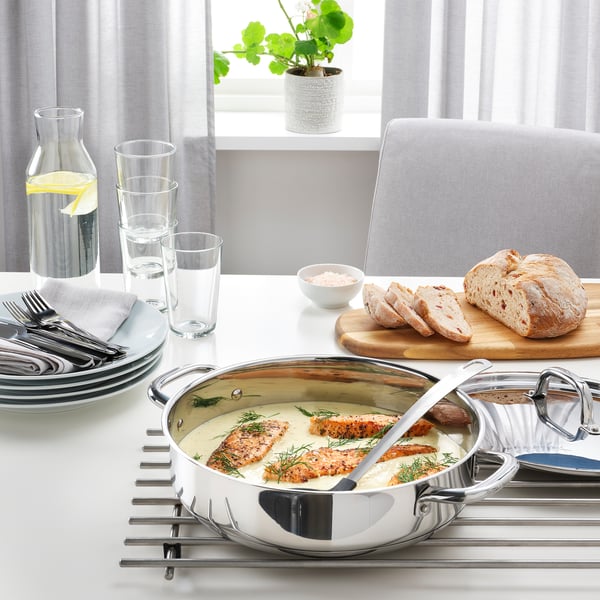 Sauterpande i rustfrit stål på bordskåner med grillet fisk, brød og glas på spisebordet ved vinduet.