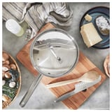 IKEA 365+ sauterpande i rustfrit stål med låg på træplade. I nærheden, svampe, kniv, ost og klud.
