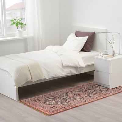 Lyst soveværelse med sprødt hvidt sengetøj, små lilla puder, ISGRÄS tæppe, hvidt natbord og lampe og vinduesplante.