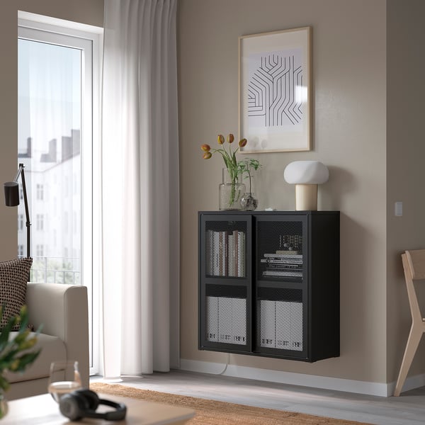 Et lille sort IVAR-netskab hænger på væggen ved siden af vinduet, med hvid lampe og orange blomster under abstrakt vægkunst.