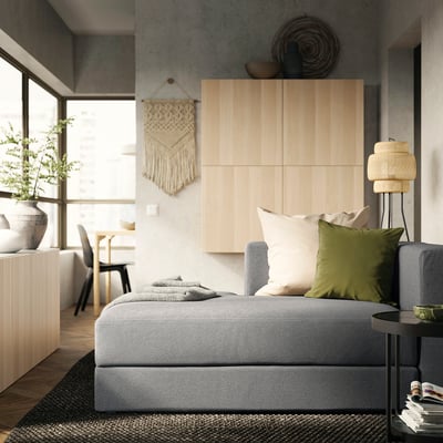 Moderne stue med grå JÄTTEBO sofa, grønne puder, vævet vægkunst, træskab og stort vindue.