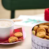 Et hvidt kaffekrus og dåse ingefærkiks på et bord, ledsaget af en rød serviet.