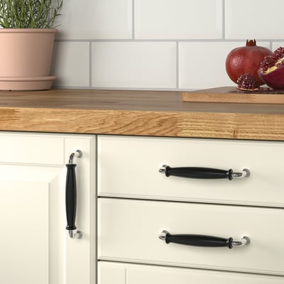 Køkkenskuffer med sorte porcelænsknopper, hvide skabe, træbordplade, potteplante og friske granatæbler.
