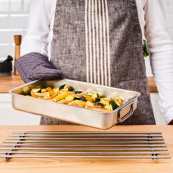 En person i et forklæde holder en bageform med grillede grøntsager over et bord med metalgrillstativer.