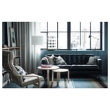 Moderne stue med store vinduer, en sort lædersofa, blåstribet tæppe, sidebord i træ, grå lænestol, gulvlampe og udsigt over byen.