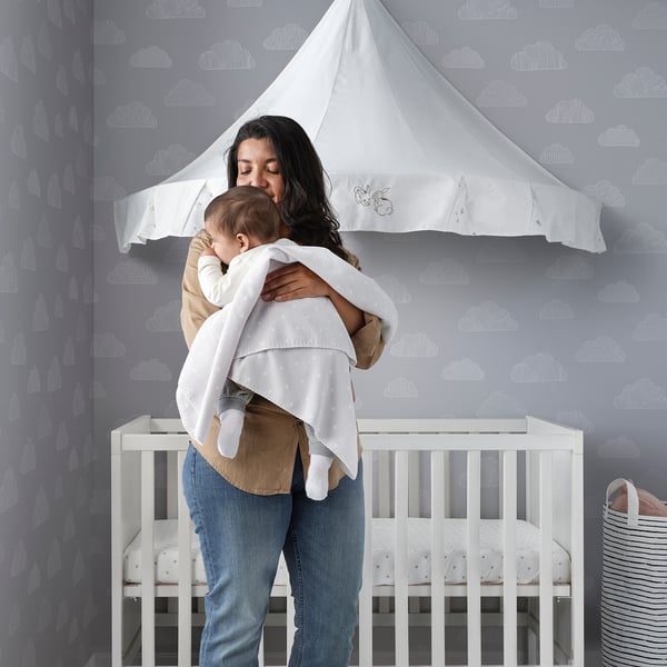 Kvinde holder baby ved vugge med skytapet og baldakin. Baby svøbt i tæppe af genanvendt polyester.