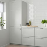 Moderne lysegråt LERHYTTAN køkken med skabe i massivt træ, planter og bagplade. Høje enheder tårner sig op over disken med flaske og redskaber.
