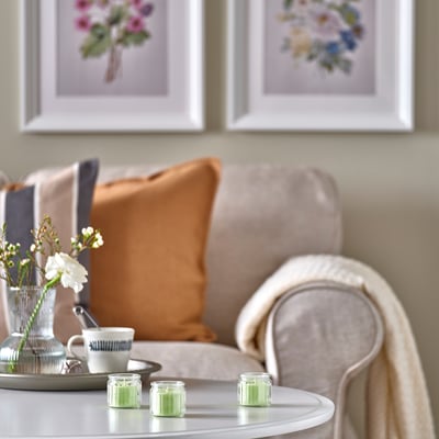 Tre grønne stearinlys på bordet ved siden af sofaen med puder, blomster og prints.
