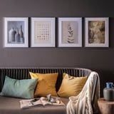 Fire indrammede kunstværker over sofaen med gule/grønne puder, hvidt tæppe, bog og vase med frugt.
