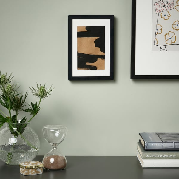 Den stilrene ramme rummer abstrakt kunst, placeret mellem en vase med grønne planter og et timeglas.