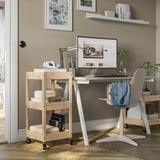 Hjemmekontor med trævogn, skrivebord og skærm. Lyse vægge, fyrretræsvogn med hylder. Grå stol med jakke.
