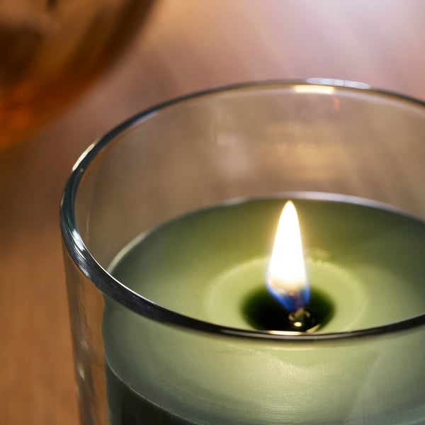 Et tændt grønt duftlys i en klar glasholder, der udsendte en klar flamme, stod på en træoverflade.