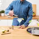 Person hælder drink fra shaker i martiniglas med jigger og lime. Tælleren viser cocktailværktøjer og ingredienser.