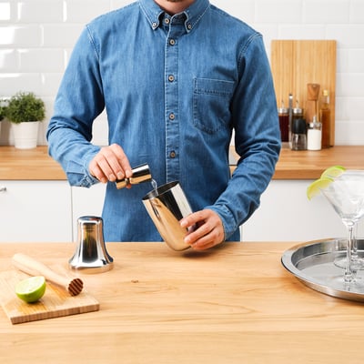 Mand hælder op fra en cocktailshaker. På træbord med limefrugter, glas og træredskaber.