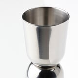Skinnende cocktailshaker i rustfrit stål, poleret overflade, der reflekterer lys, står alene på en reflekterende overflade.