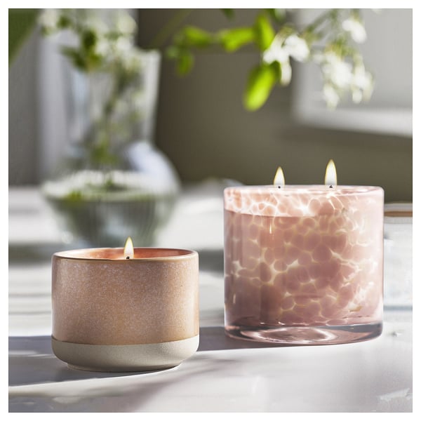 To stearinlys brænder: beige i keramik, lyserød med honeycomb i glas. I nærheden af vindue med planter.