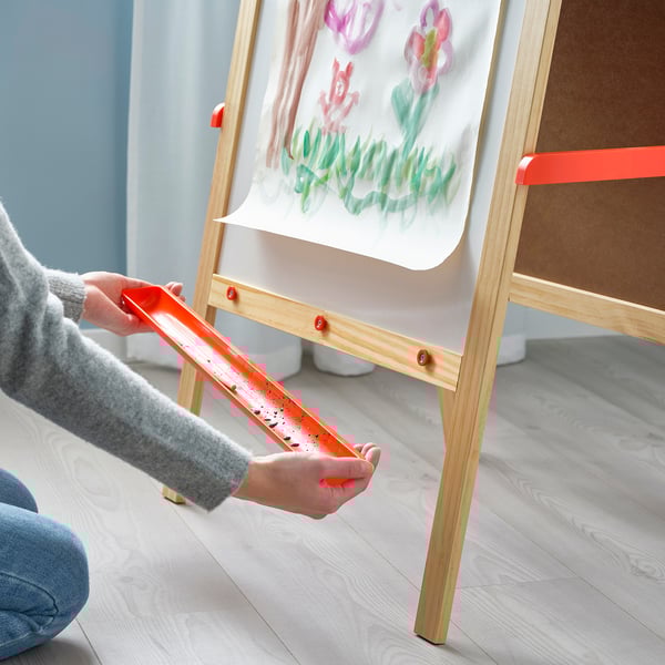 Børnehænder holder en orange legetøjssaks, klippepapir på et træstaffeli med en tegning af blomster.