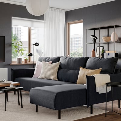 Moderne stue med MANNARP grå sofa, store puder, sideborde, hylder og store vinduer med hvide gardiner.