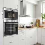 Moderne køkken med hvide skabe, hvidevarer i rustfrit stål, bordplade i træ med skærebræt og tomater.