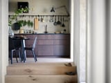 Moderne køkken med mørkebrune skabe og trækanter, hængende redskaber og et sort spisebord med stole.