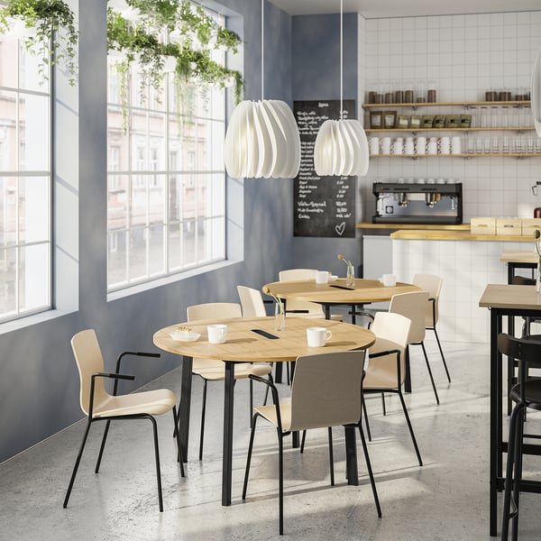 Moderne café med træborde, hvide stole, grønne planter og store vinduer.