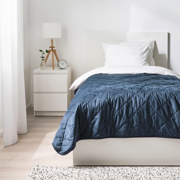 Minimalistisk soveværelse: hvid seng med mørkeblå MJUKPLISTER genbrugsfløjlsdyne, matchende hvide puder, lille natbord med lampe og ur.
