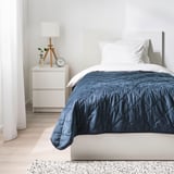 Minimalistisk soveværelse: hvid seng med mørkeblå MJUKPLISTER genbrugsfløjlsdyne, matchende hvide puder, lille natbord med lampe og ur.