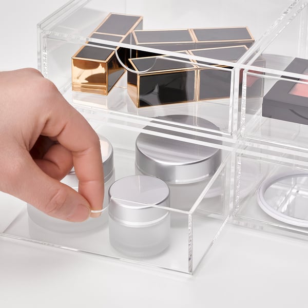 Hånden rækker ind i klare, stabelbare akrylopbevaringsskuffer og organiserer makeup og smykker.