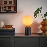 Orange MOLNART pære i sort marmorfod, med gylden skål, hvid kugle, plante og billede.