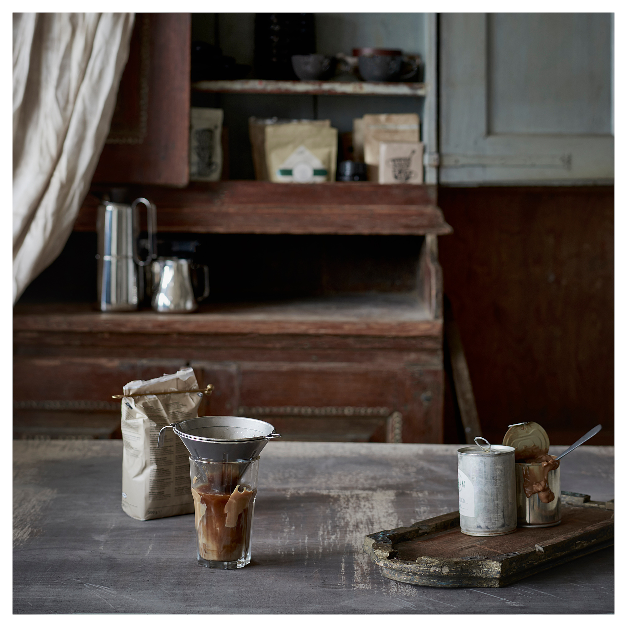 Vintage køkken med kaffeopsætning, herunder et sølv kaffefilter, bønner i poser og beholdere med redskaber på et rustikt træbord.