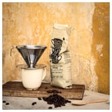En kaffescene med et genanvendeligt sølvfilter, hvid kop og brune bønner. Taskelæsninger nyder øjeblikket med en gul baggrund.