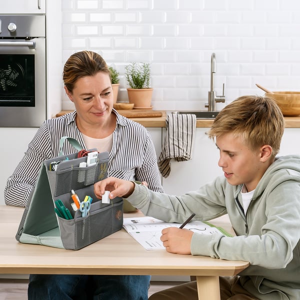 En kvinde hjælper en dreng med hans lektier ved et køkkenbord ved hjælp af en ÖVNING-arrangør, som indeholder skoleartikler og en tablet.
