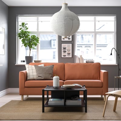 Moderne stue med store vinduer, mørke vægge, orangestribet sofa, pendel, sofabord med bøger, skammel og plante.