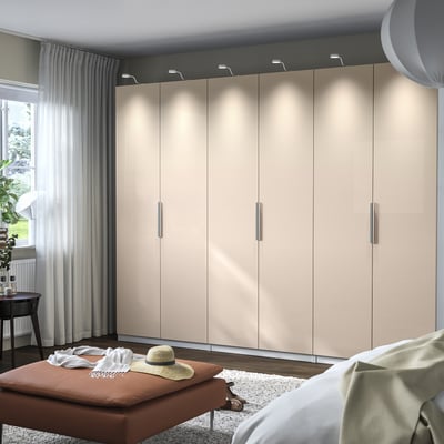 Et stort, beige PAX garderobeskab med soft-close hængsler. Garderobeskabet er oplyst og har et slankt, moderne design, der står højere end møbler i nærheden.