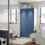 Moderne soveværelse med blå PAX garderobe og hvid stol. Garderobeskabet højt, bredt, med sølvhåndtag. Stort vindue ved siden af.