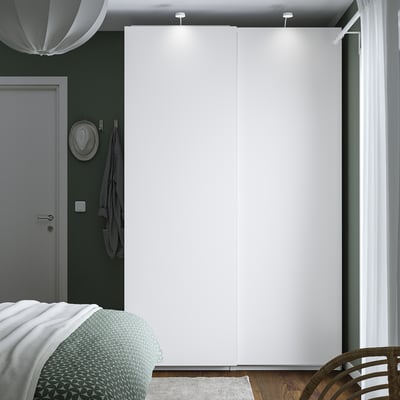 Minimalistisk soveværelse med stort PAX garderobeskab, slanke døre og belysning. Grå dør, grønt sengetæppe, hvide gardiner, hat og håndklæde ved dør.