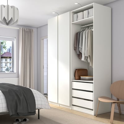 Moderne hvid PAX garderobe tårner ved siden af sengen og opbevarer tøj, tasker og kasser. Høj, med skuffer i bunden og hængeskinne.