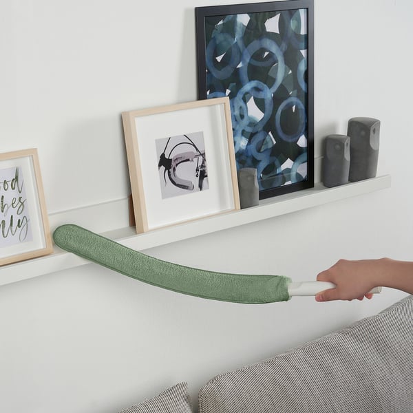 En hånd, der holder en grøn PEPPRIG mikrofiber-støvsuger og strækker den vandret over en hvid hylde med vægdekoration.