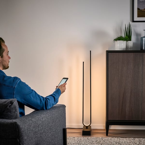 Mand sidder på sofaen ved hjælp af smartphone, sandsynligvis styrer moderne gulvlampe med trådløs lysdæmper. Lampen har et unikt, bøjet led design.