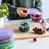 En person placerer låget på en beholder med druer. På en træplade er der flere druer og silikonebeholdere med låg til opbevaring af mad.