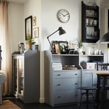 Moderne stue med grå skabe, et rundt vægur, sort lampe og dekorative genstande med spisebord og stole i baggrunden.