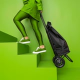 En person iført grønt sportstøj og hvide sneakers skubber en sort indkøbsvogn op ad grønne trin.