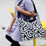 Person med sort og hvid RÄCKLA skuldertaske, større end en vandflaske, i abstrakt mønster, iført lyselilla top og sorte leggings.