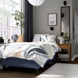 Moderne soveværelse med RÅHOLMAR seng, gråhvid dyne, natbord i træ med lampe, ur og plante ved vindue.