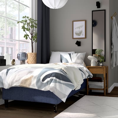 Moderne soveværelse med RÅHOLMAR seng, gråt sengetøj, natbord i træ, lampe, ur og plante.