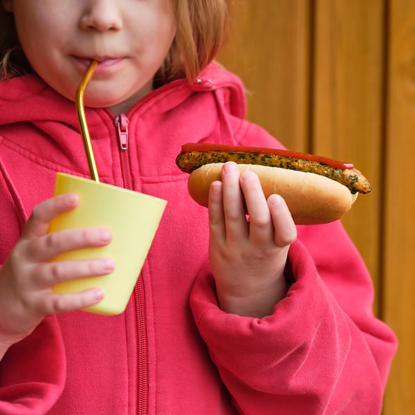 Barn i rød hættetrøje drikker fra gul kop, holder grillet hotdog.