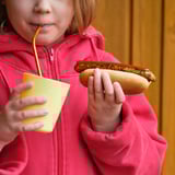 Barn i rød hættetrøje drikker fra gul kop, holder grillet hotdog.