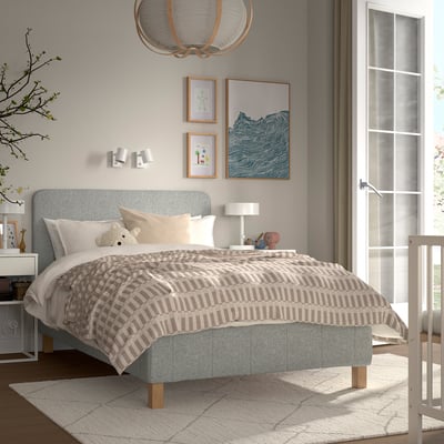 Moderne soveværelse med grå seng, ternet dyne, puder, bamse, kunst, natbord med lampe og plante, delvis krybbe.