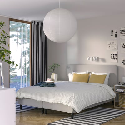 Moderne soveværelse med RENFJÄLLET-seng , grå sengegærde, hvid lampe, stort vindue, stribet tæppe, gul pude, potteplante.